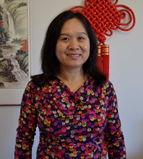A woman with dark hair stands and smiles at the camera. She is wearing a colorful, floral-patterned dress. Behind her, there is a traditional red Chinese knot decoration on the wall and a framed painting with nature scenery.