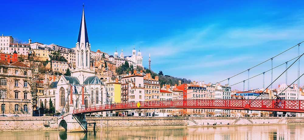 A scenic view of a European city featuring a red bridge spanning a river. In the background, there's a mixture of historic buildings with red roofs, and a prominent church with a tall spire. The sky is clear and blue, adding to the vibrant atmosphere.