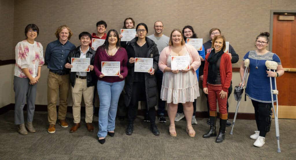 A group of thirteen people, some holding certificates, stand together and smile in a room with beige walls. One person on the right uses crutches and has a leg cast. They appear to be celebrating an achievement.
