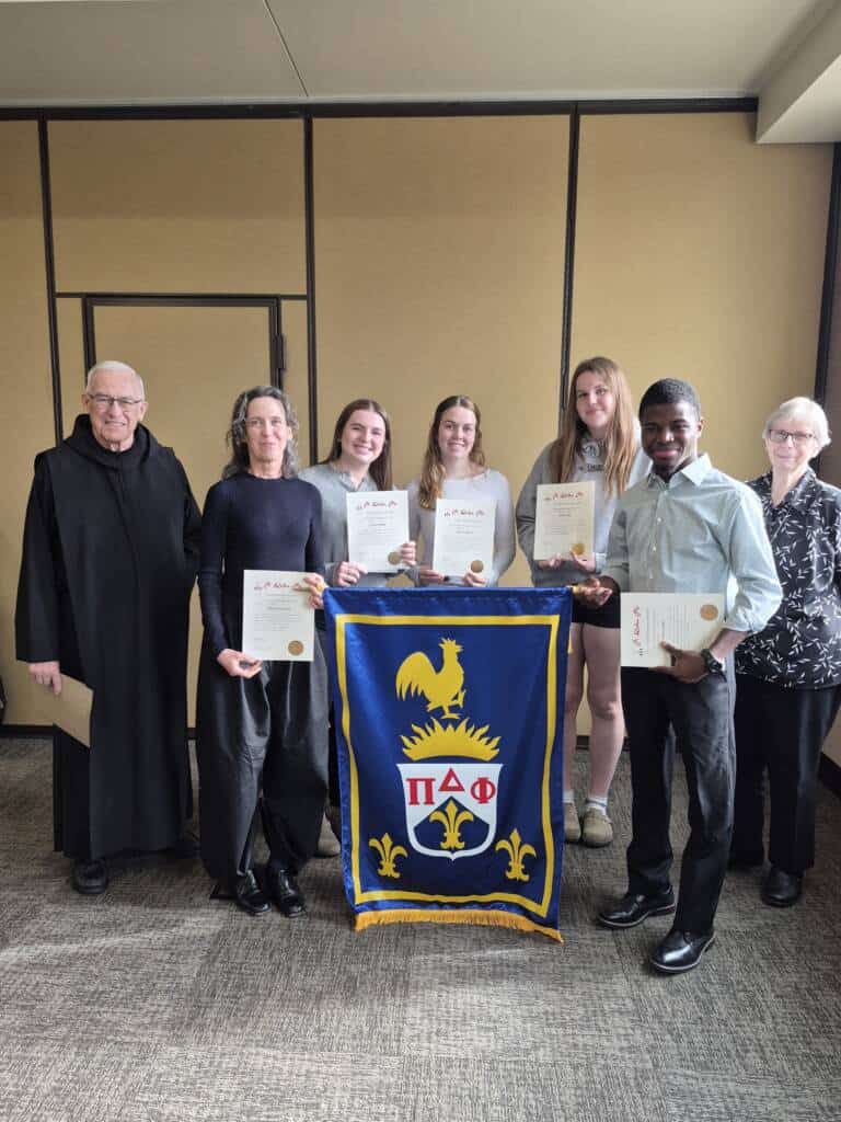 Six people stand indoors, smiling and holding certificates. Four young people stand in the center, holding a blue and gold banner with a rooster and Greek letters. Two older adults stand at each end, one in a black robe and the other in a white blouse.