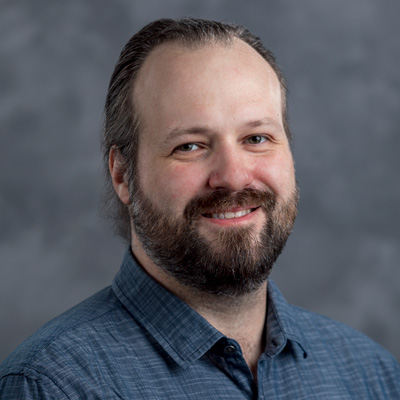 A bearded man with long hair smiles at the camera. He is wearing a blue button-up shirt. The background is a gray gradient.