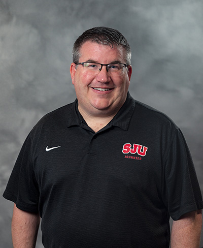 A smiling man wearing glasses and a black polo shirt with a red "SJU Johnnies" logo stands against a gray, textured background.