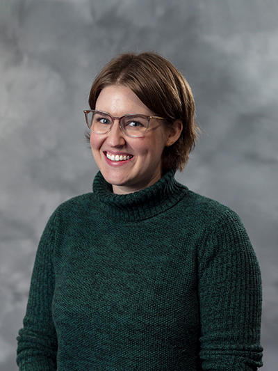 A person with short brown hair and glasses smiles at the camera. They are wearing a dark green turtleneck sweater. The background is a gray, textured wall.
