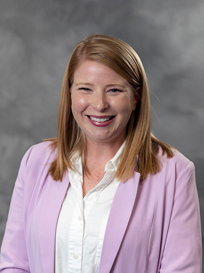 A smiling person with straight, shoulder-length hair, wearing a light pink blazer over a white shirt. The background is a neutral gray.