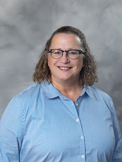 A person with curly hair and glasses smiles while wearing a blue and white striped shirt. The background is a soft gray gradient.