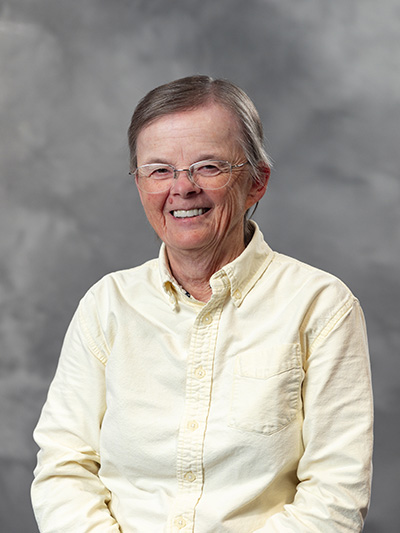 An elderly person with short hair and glasses smiles warmly, wearing a light-colored shirt against a gray backdrop.