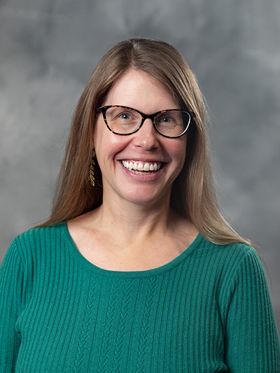 A woman with long brown hair and glasses is smiling and wearing a green sweater. The background is a plain gray.