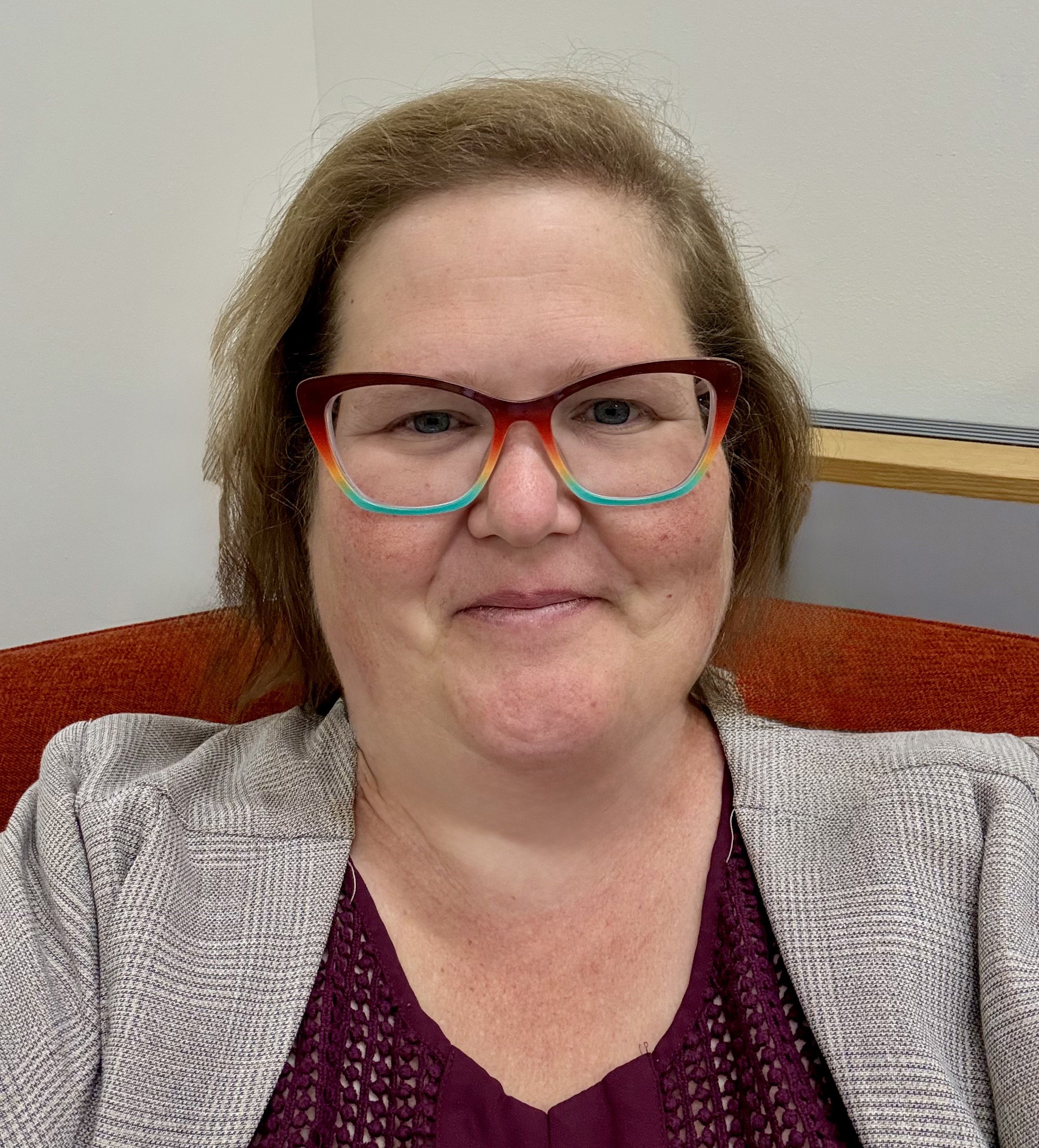 A woman with light brown hair, wearing red and teal glasses, a gray blazer, and a purple top, smiles while sitting on an orange chair in a room with white walls.