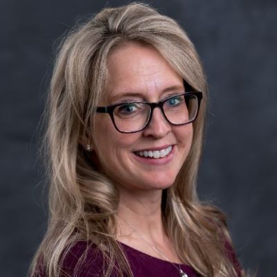 A woman with long blonde hair and glasses smiles at the camera. She is wearing a dark purple top against a dark gray background.