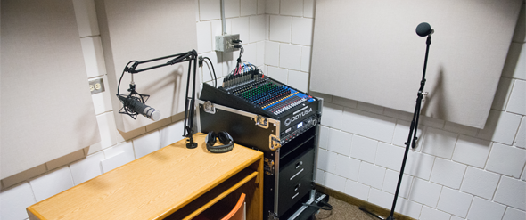 A small recording studio with a microphone on a boom arm over a desk, headphones, a mixing console with numerous controls, and a standing microphone. The walls are covered with acoustic panels.