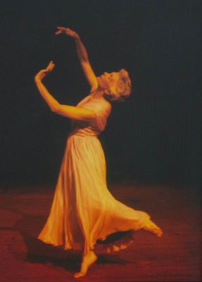 A dancer in a flowing dress performs gracefully on stage, illuminated by warm, dramatic lighting. Her arms are elegantly extended upwards, and her dress swirls gently, capturing a sense of motion and poise.