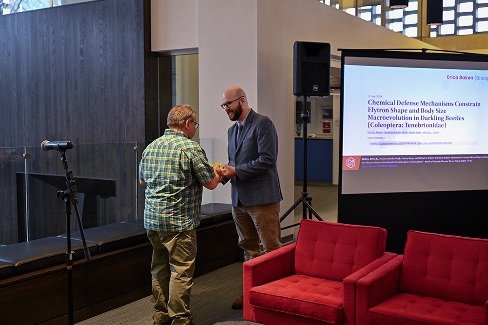 Two people stand shaking hands in front of a presentation screen displaying a scientific talk. One person is handing an object to the other. Red chairs are in the foreground, and the setting appears to be a lecture hall or conference room.