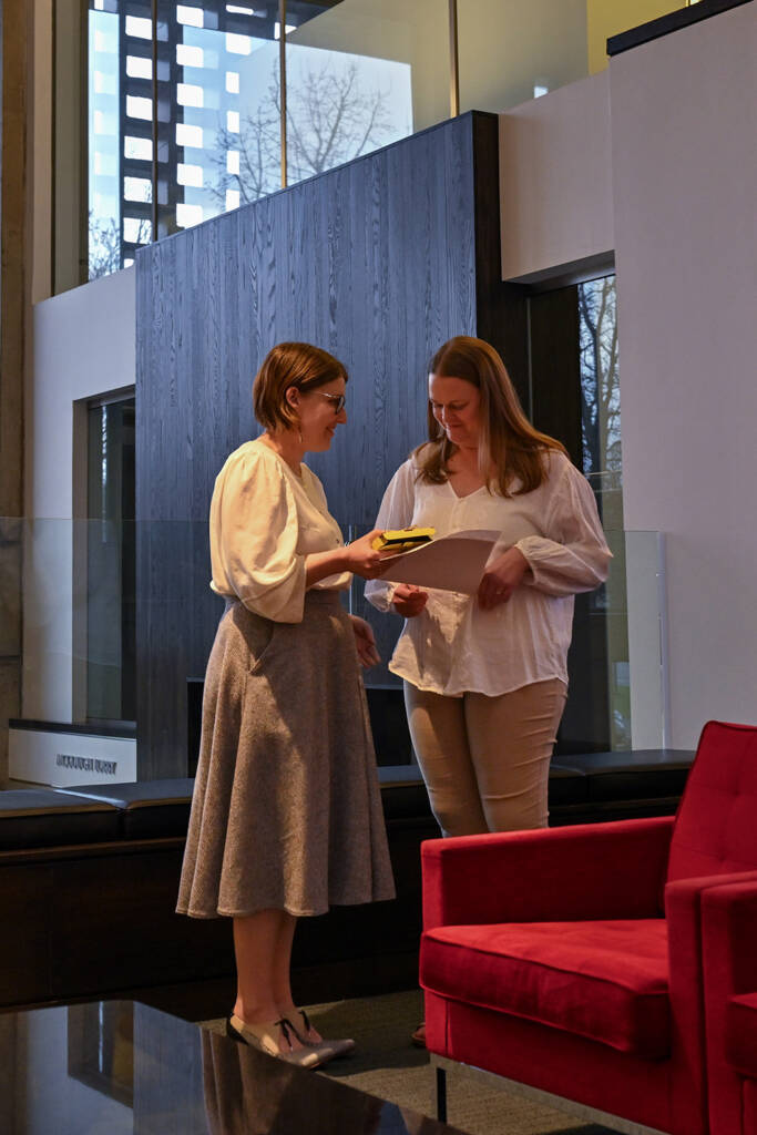 Two women stand indoors by a red chair, looking at papers together. Both wear light-colored tops; one holds a notepad, the other holds a folder. Large windows and modern decor are visible in the background.