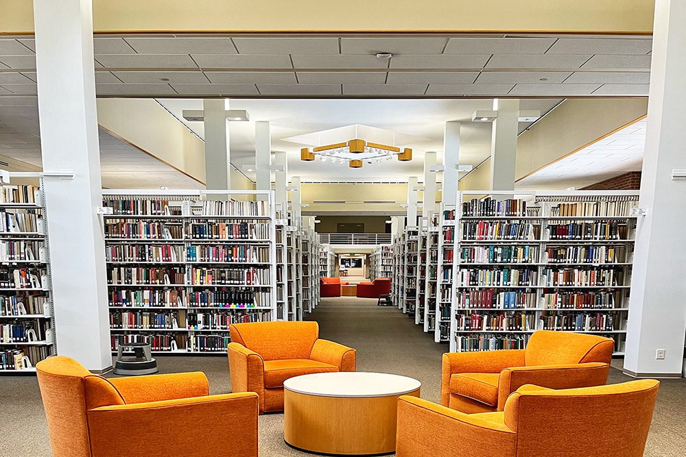 A library interior with tall shelves full of books, four bright orange armchairs around a small round table in the foreground, and more seating visible in the distance.