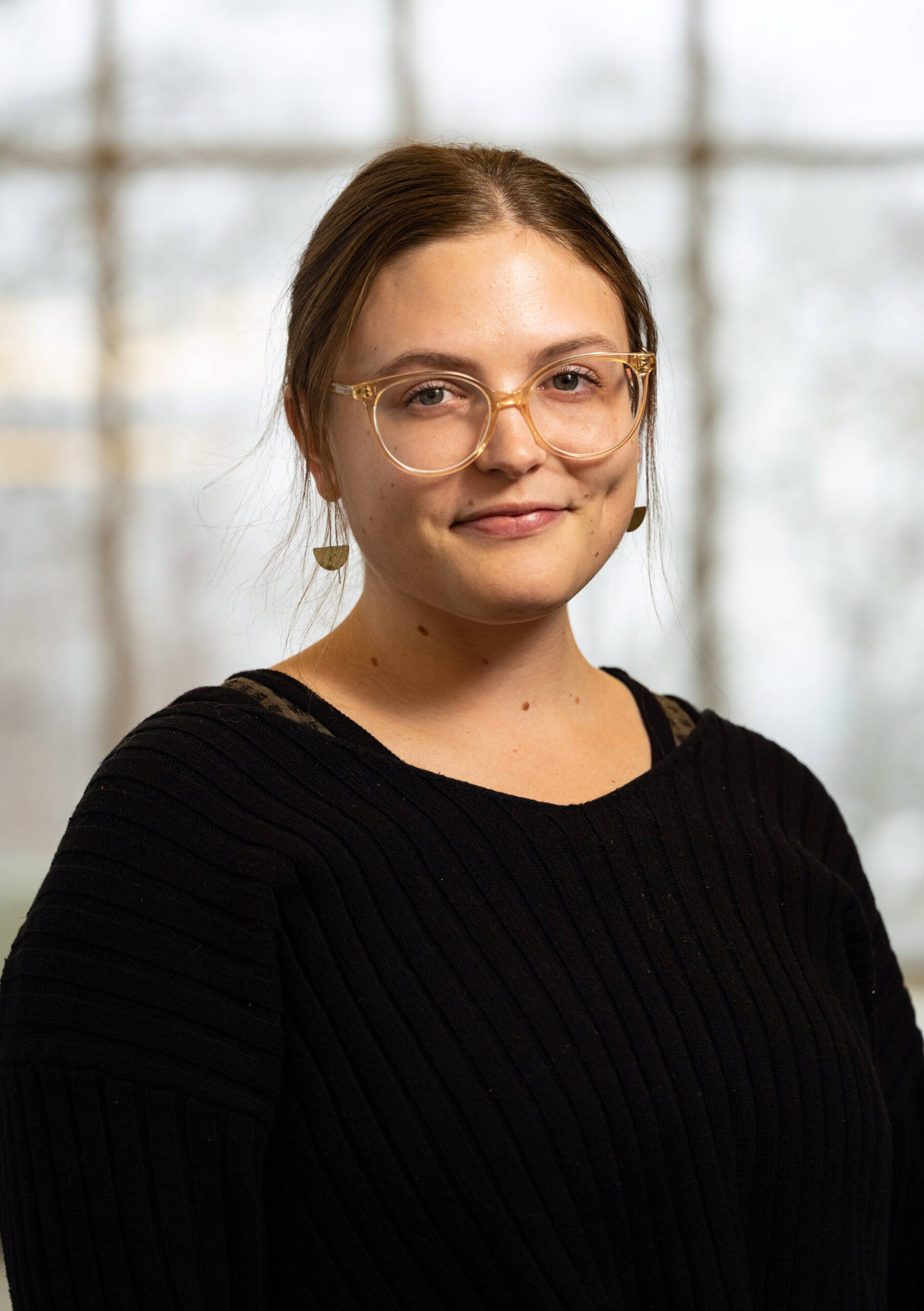 A woman with light brown hair tied back, wearing clear glasses, gold earrings, and a black ribbed sweater, stands in front of a blurred window background, smiling slightly.