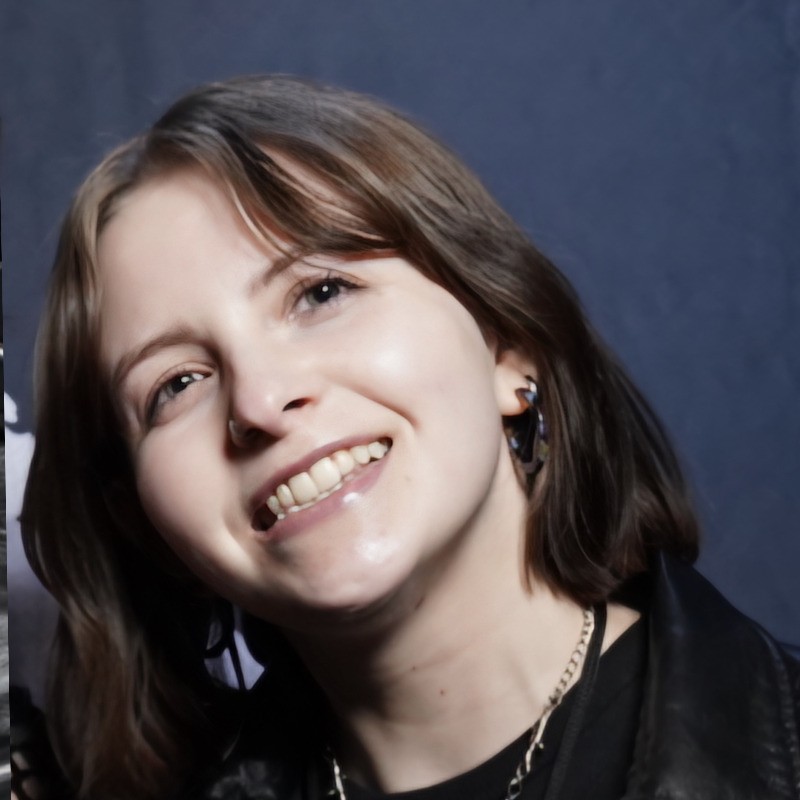 A young woman with short brown hair smiles while looking slightly upward. She is wearing a black jacket, a dark shirt, earrings, and a silver chain necklace. The background is a plain dark blue.