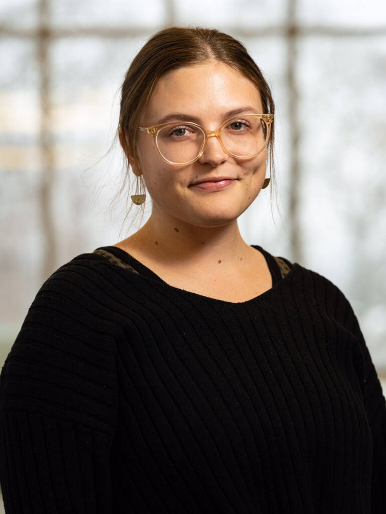 A young woman with light brown hair, wearing clear-framed glasses and a black sweater, stands indoors in front of a blurry window, smiling slightly at the camera.