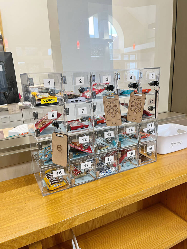 A transparent locker box with 20 numbered compartments, each holding keys with tags and some tools, sits on a wooden counter near a glass window in a public or office setting.