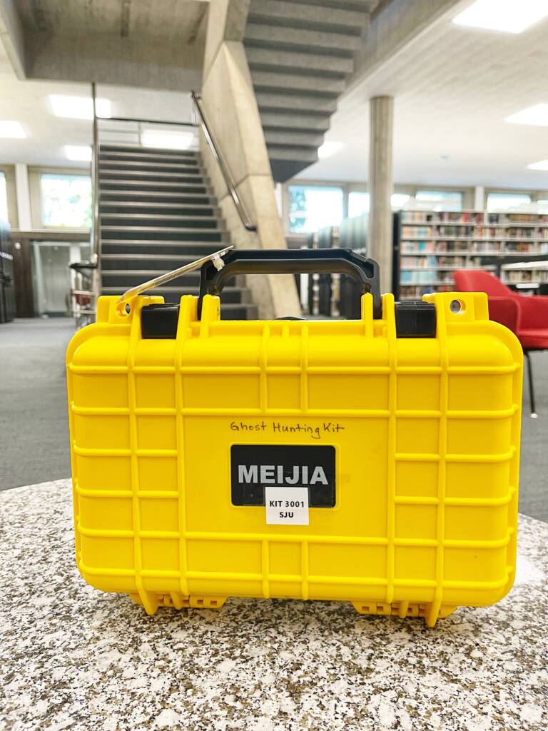 A bright yellow case labeled "MEIJIA" and "Ghost Hunting Kit" sits on a speckled surface in a library, with bookshelves and a staircase in the background.