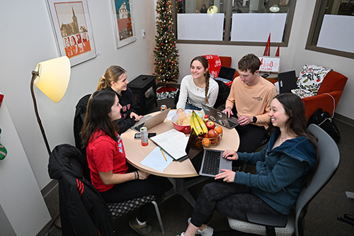 Five college students sit around a small round table studying with laptops and notebooks in a cozy room decorated with a Christmas tree and holiday decor. They are smiling and talking together.