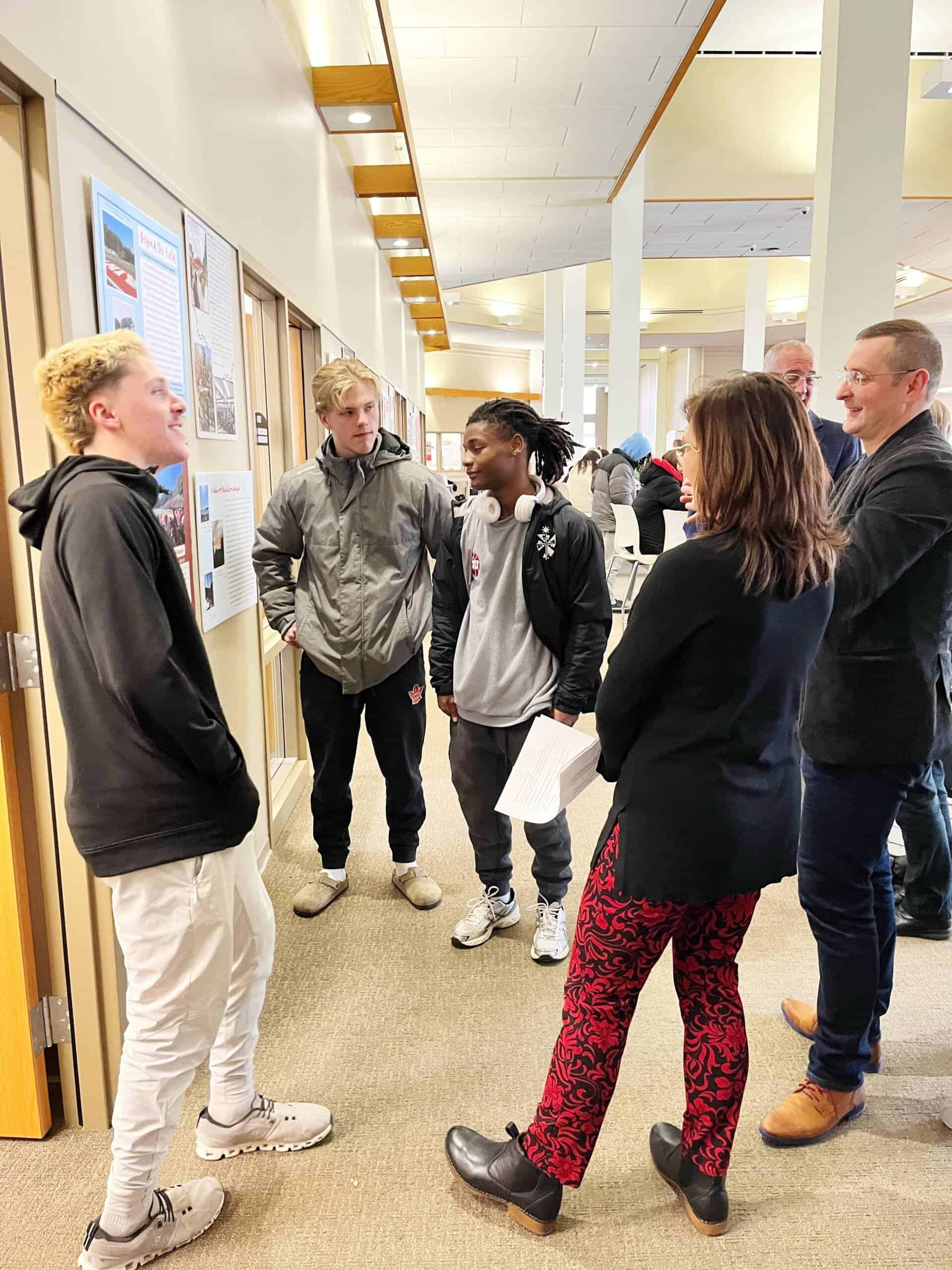 A group of three students stands presenting their work on a wall to two adults in a brightly lit hallway. The group appears engaged in conversation, with others visible in the background.