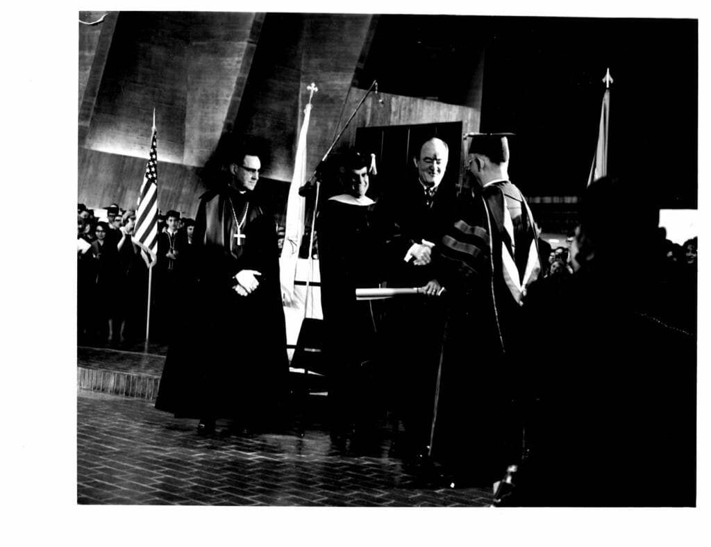 A graduation ceremony with graduates in caps and gowns receiving diplomas on stage; officials in academic regalia stand nearby. American flags are visible in the background.