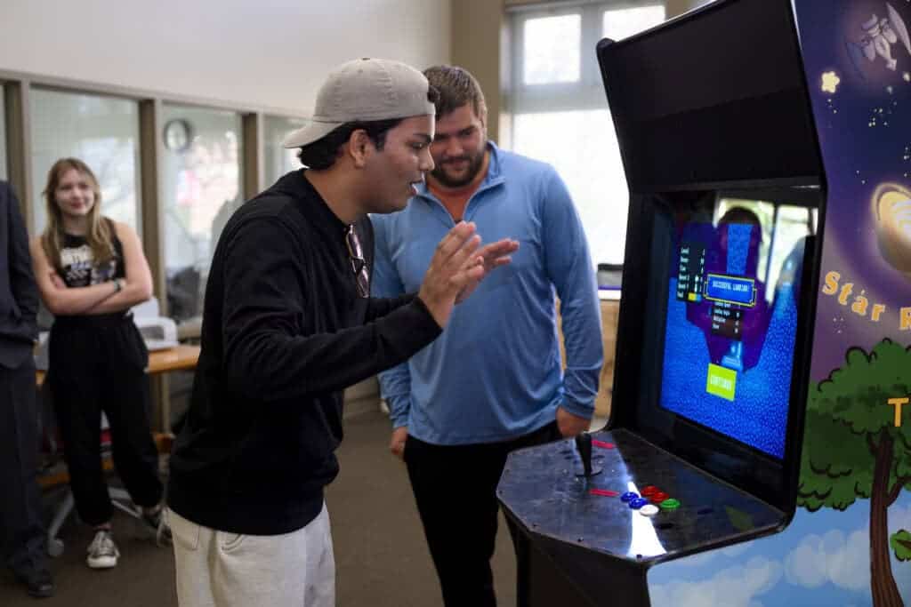 A man wearing a cap excitedly plays an arcade game while another man in a blue shirt watches and smiles. Two people stand in the background near a window, observing the scene.