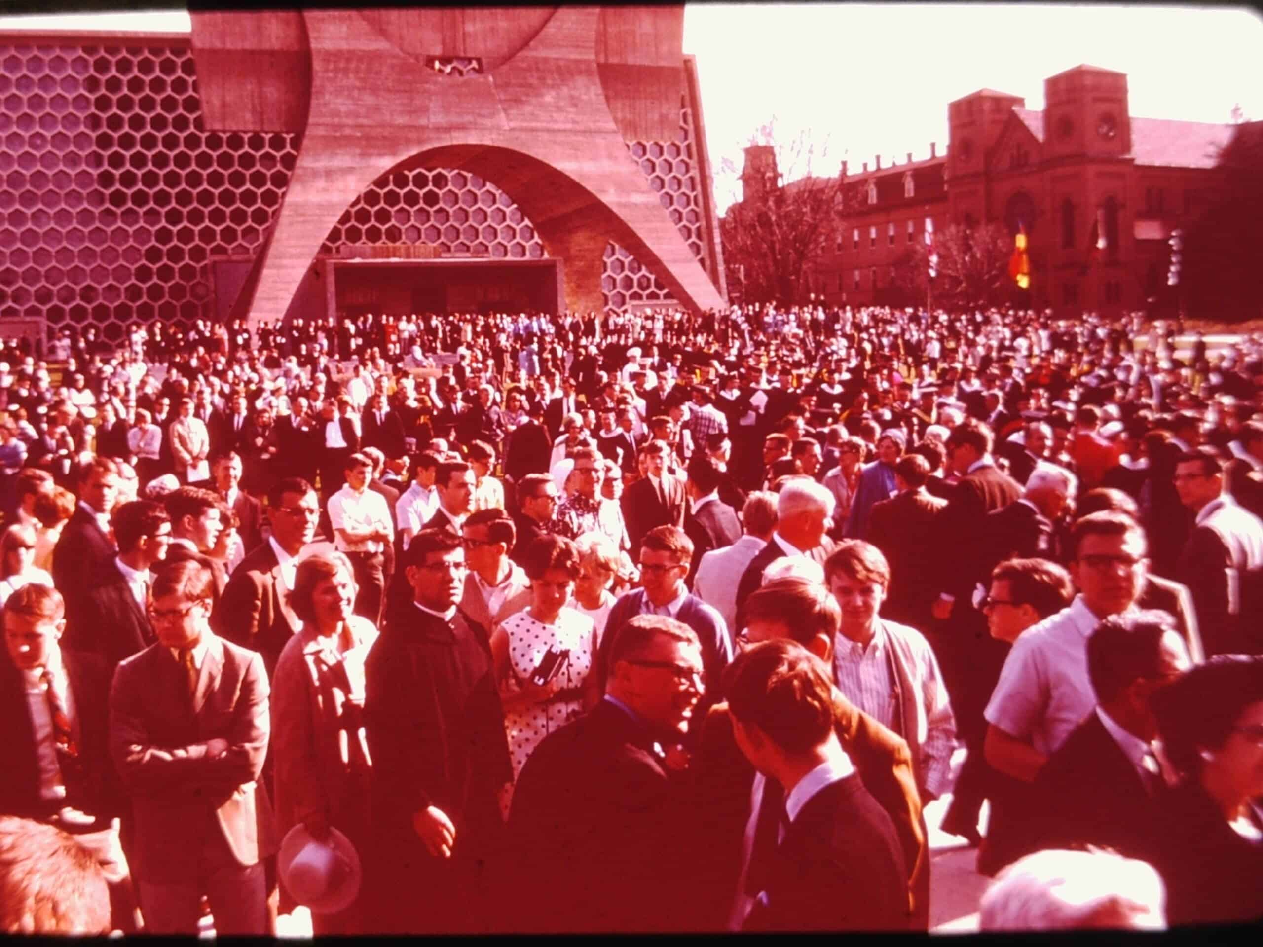 A large crowd of people, many dressed in suits and dresses, gathers outside a modern honeycomb-patterned building on a sunny day. Older brick buildings and trees are visible in the background.