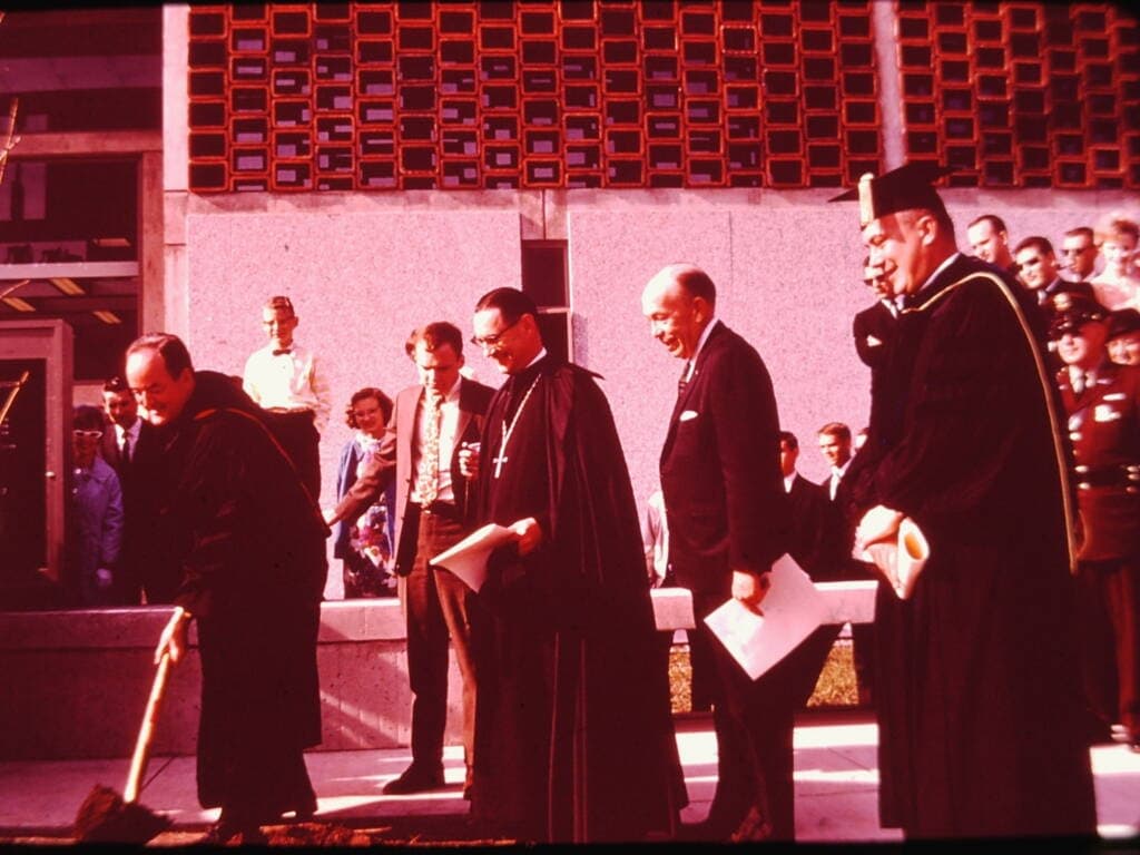 Several men in religious and academic robes participate in a ceremonial groundbreaking event outside a modern building, while a crowd watches in the background. One man is using a shovel to break ground.