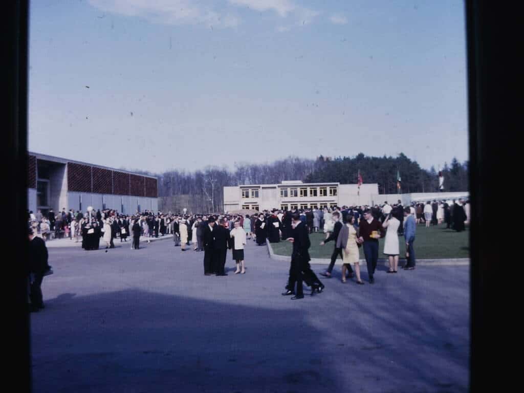 A large group of people, many dressed formally, gather on a paved area and lawn outside modern buildings on a sunny day, with trees and a blue sky in the background.