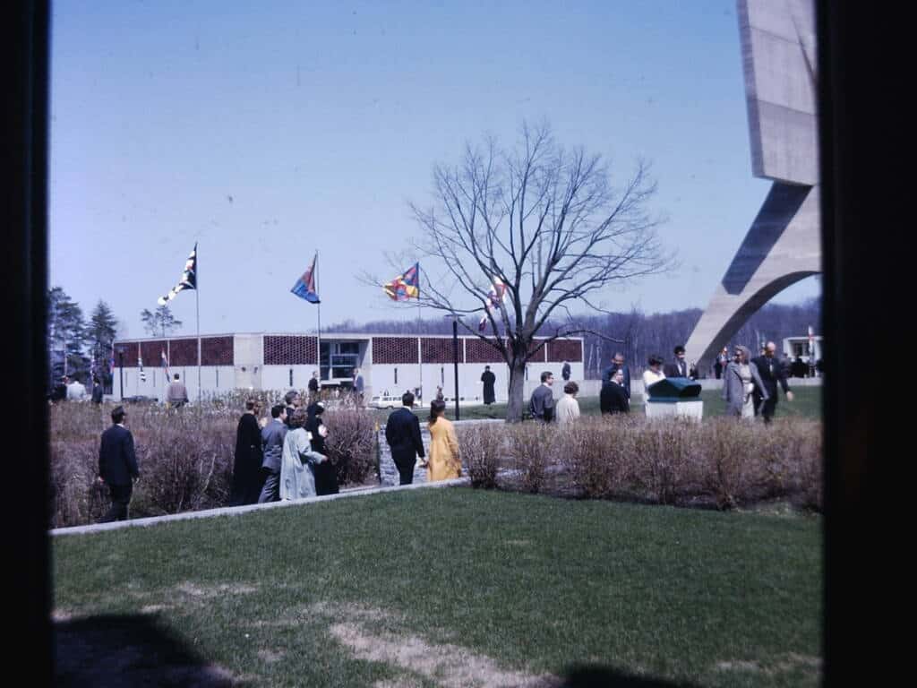 A group of people walks through a grassy area with bushes toward a modern concrete structure. Several colorful flags are displayed near a leafless tree under a clear blue sky.