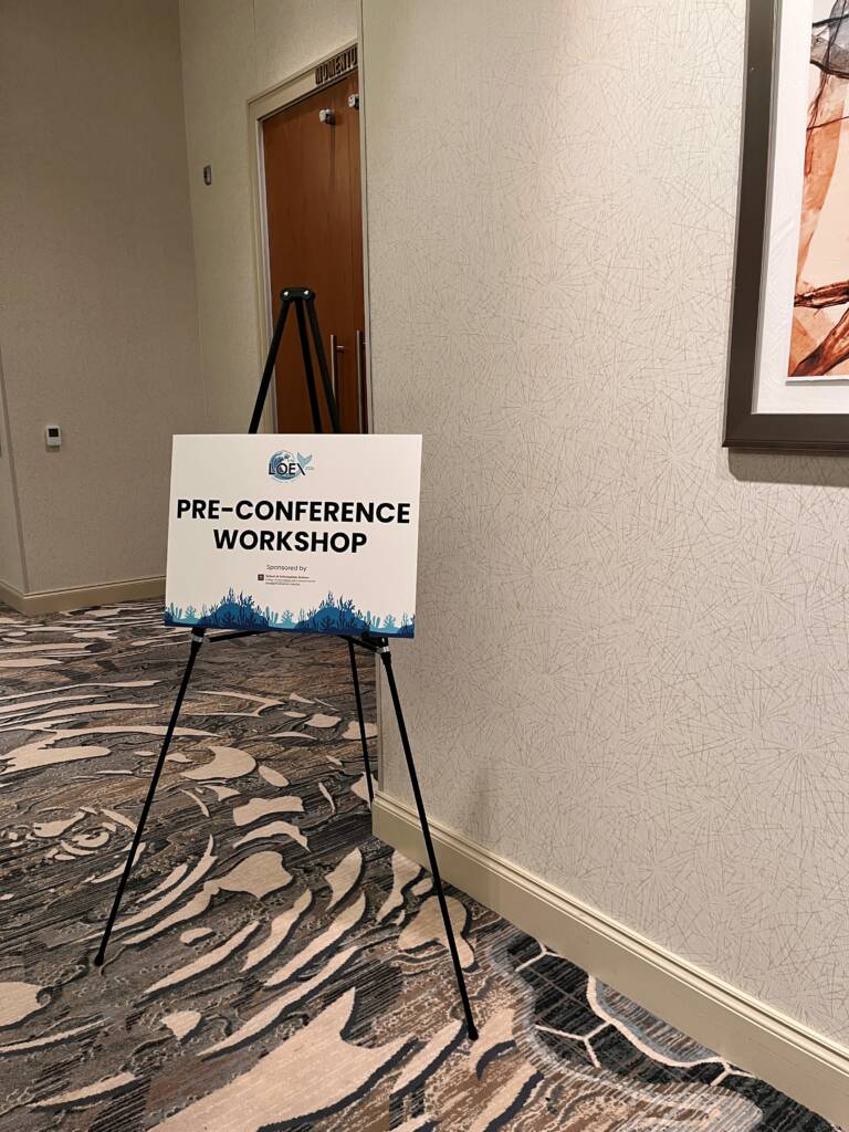 A sign on a tripod stand in a hallway reads "Pre-Conference Workshop" with a conference logo above. The sign is next to a closed door and abstract artwork on the wall.