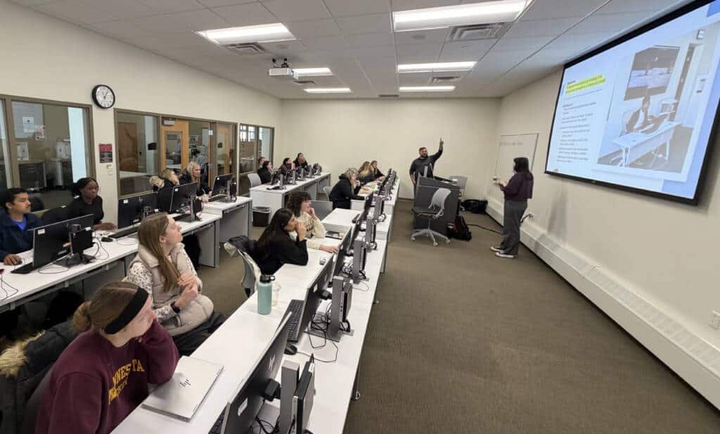 Students sit at rows of desks with computers in a classroom, attentively watching two people present slides on a large screen at the front of the room. One presenter points at the screen while the other stands nearby.