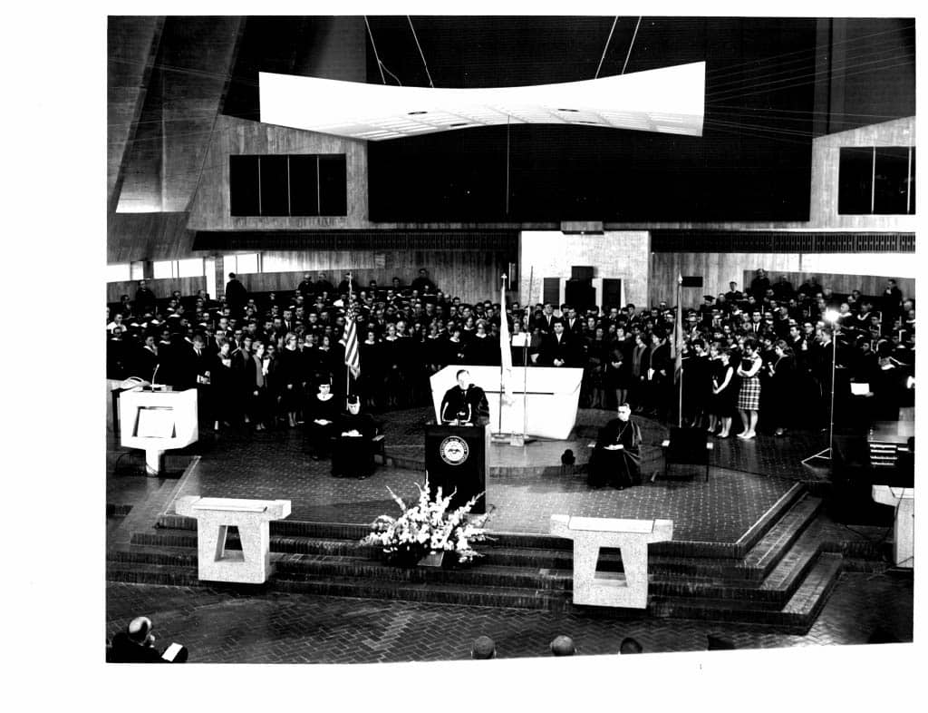 A large group of people gathered inside a modern church or auditorium, watching a person speak at a podium. Several flags and flower arrangements are present, with two seated figures in front of the audience.