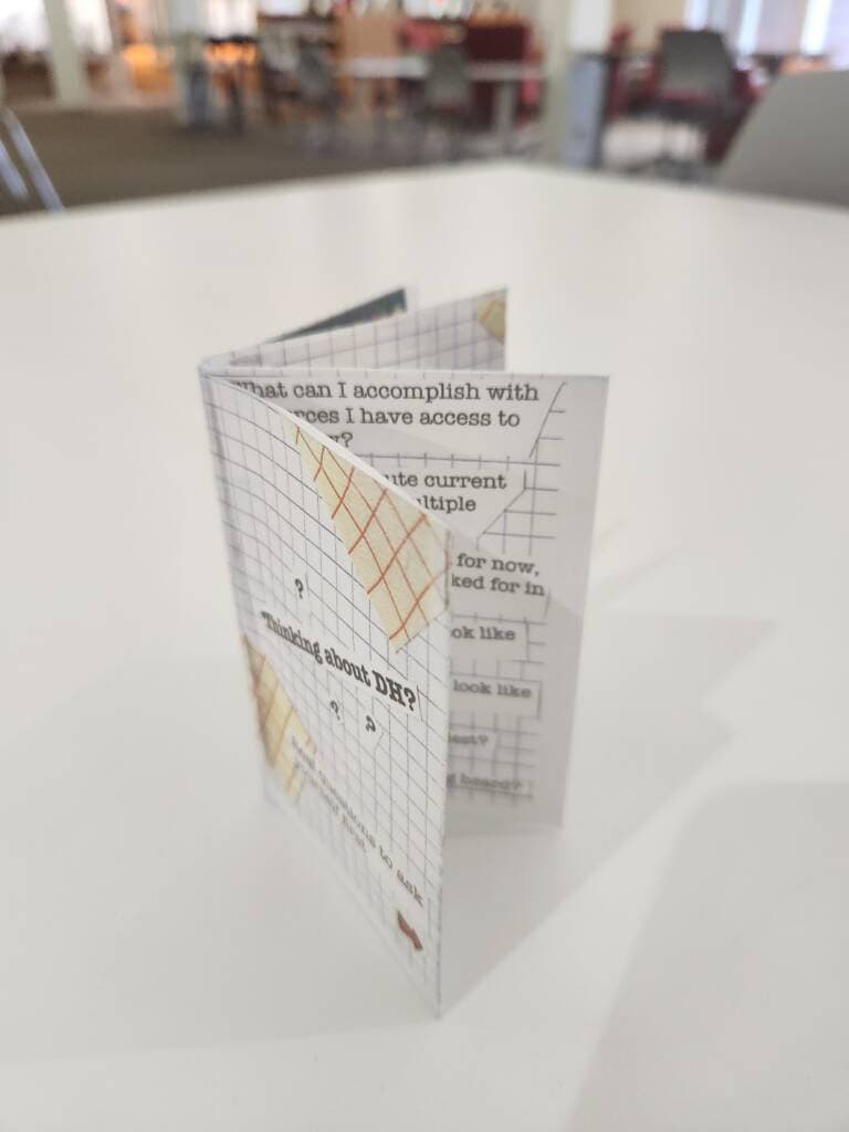 A small, handmade booklet with a grid design and partially visible printed text stands upright on a white table in a brightly lit room with blurred chairs and tables in the background.