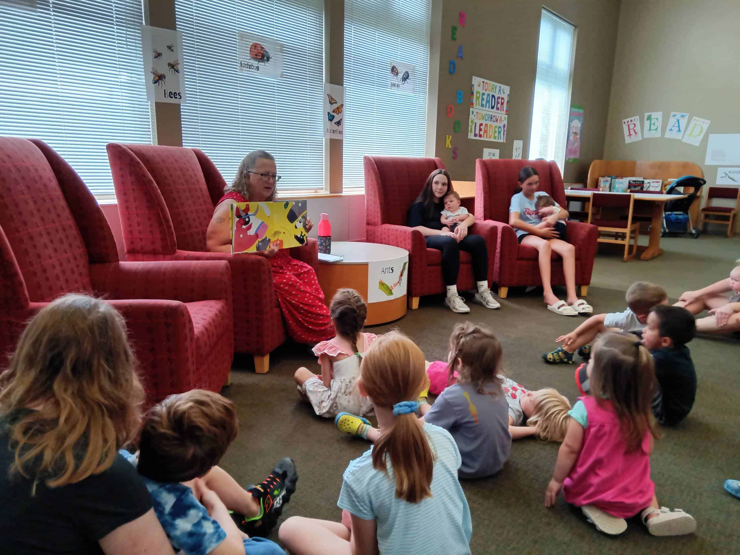 A woman sits in a red chair reading a picture book to a group of young children seated on the floor in a library, while two older children listen from nearby chairs. Bright posters and bookshelves decorate the room.