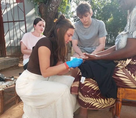 A young woman wearing blue gloves examines the hand of an older person seated outdoors, while two other young people observe nearby. They are engaged in an activity, possibly medical or educational, in a rural setting.