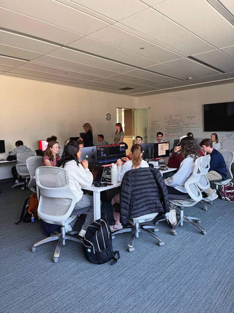 A group of students sit around tables with computers in a modern classroom, collaborating and working. Some are talking, while others focus on their laptops; whiteboards and screens are visible in the background.