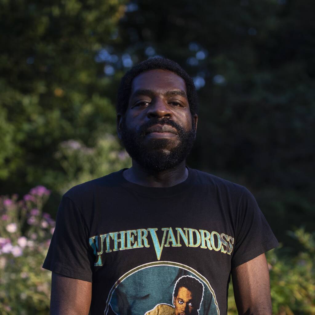 A person with short hair and a beard stands outdoors. They are wearing a black T-shirt featuring an image and the name "Luther Vandross." The background is a lush garden with various green plants and some visible flowers. The lighting is natural and bright.