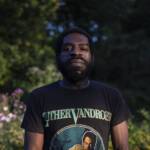A person with short hair and a beard stands outdoors. They are wearing a black T-shirt featuring an image and the name "Luther Vandross." The background is a lush garden with various green plants and some visible flowers. The lighting is natural and bright.