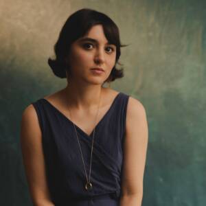 A woman with short, dark hair is looking toward the camera with a neutral expression. She is wearing a sleeveless dark blue dress and a long necklace with a circular pendant. The background is textured with shades of green and blue.