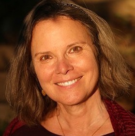 A smiling woman with shoulder-length brown hair wears a dark burgundy top and a gold earring. She is outdoors, and the warm sunlight softly illuminates her face. The background is blurred, highlighting her as the main focus of the image.