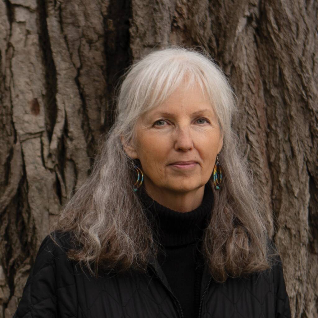 A woman with long gray hair wearing a black quilted jacket and turquoise earrings stands in front of a tree trunk. She has a gentle expression and is looking directly at the camera.