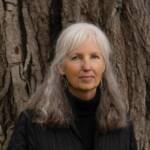 A woman with long gray hair wearing a black quilted jacket and turquoise earrings stands in front of a tree trunk. She has a gentle expression and is looking directly at the camera.