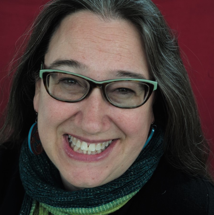 A person with long brown hair, wearing glasses and a green scarf, is smiling warmly against a reddish background. The person's expression is cheerful, and they are wearing teal earrings.