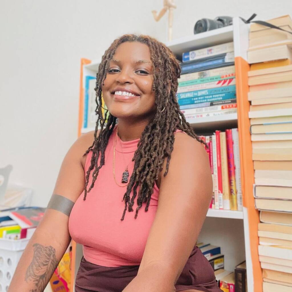 A woman with long braided hair smiles while posing in front of a filled bookshelf. She is wearing a sleeveless, peach-colored top and several necklaces. A tattoo is visible on her arm. Stacked books and other items are visible in the background.