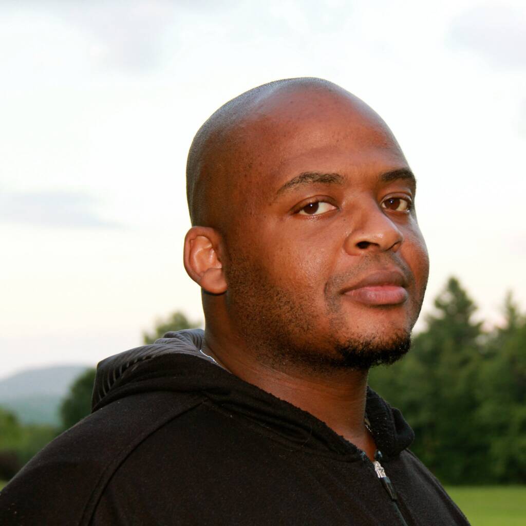 A man in a black hoodie stands outdoors with a serene landscape in the background. The scene includes a clear sky, distant hills, and lush green trees. He has a neutral expression on his face and looks directly at the camera.