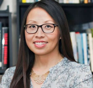 A woman with long dark hair and glasses smiles at the camera. She is wearing a patterned blouse and a gold necklace. Bookshelves filled with books are visible in the background.