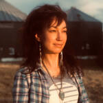 A woman with dark hair stands outdoors, looking into the distance with a thoughtful expression. She is wearing a plaid shirt over a T-shirt with the word "sunset" and beaded jewelry. Farm silos and a field are visible in the background. The sun casts a warm glow.