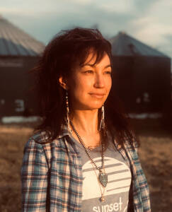 A woman with dark hair stands outdoors, looking into the distance with a thoughtful expression. She is wearing a plaid shirt over a T-shirt with the word "sunset" and beaded jewelry. Farm silos and a field are visible in the background. The sun casts a warm glow.